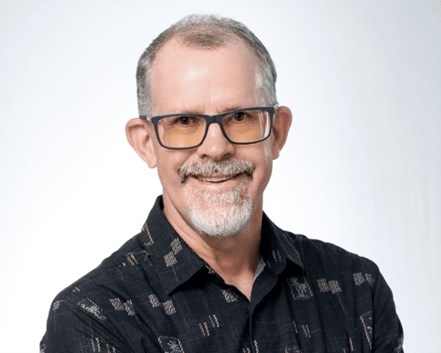 Smiling man with glasses and a beard, wearing a black patterned shirt.